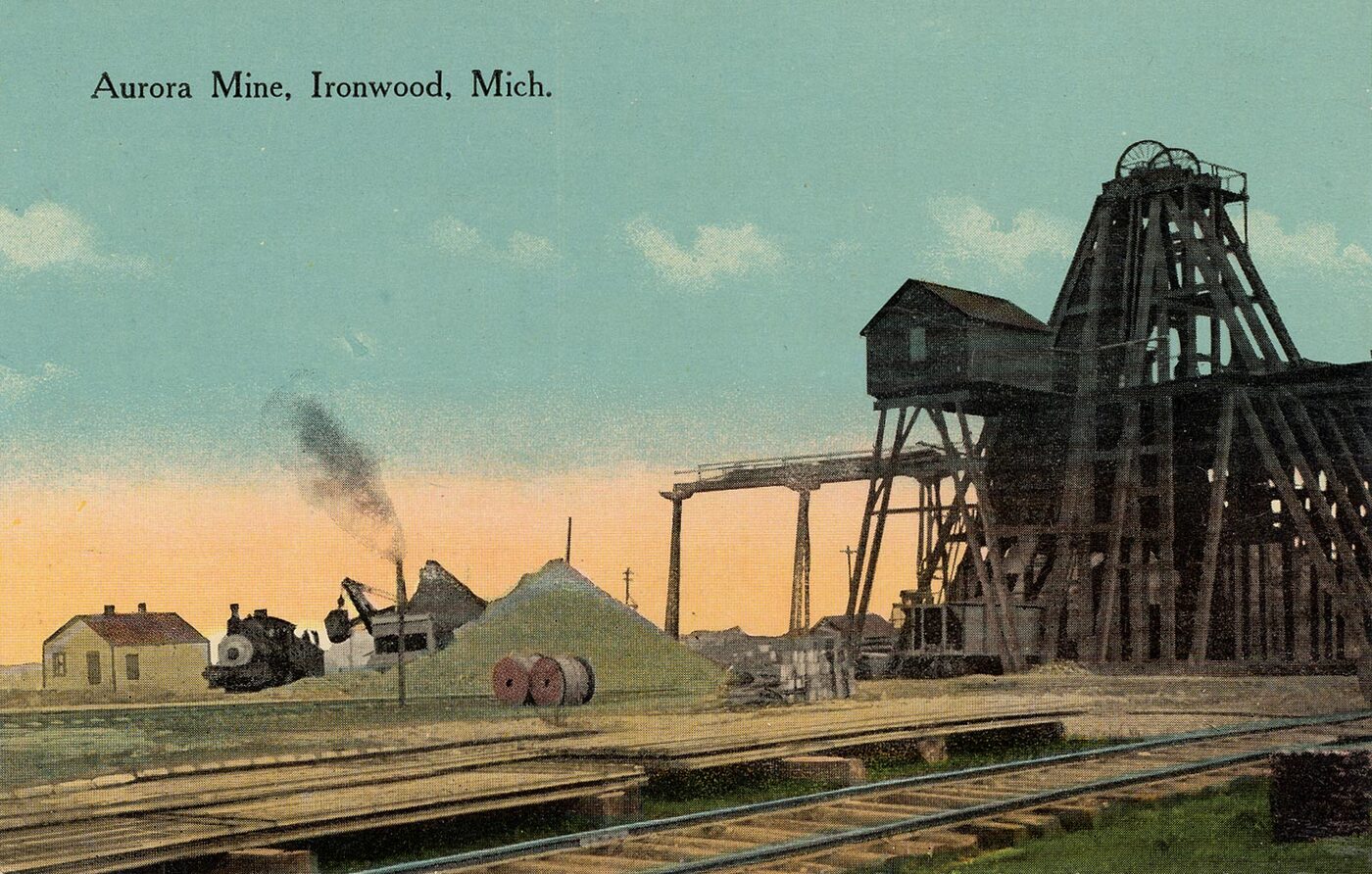 Color postcard of the Aurora Mine: a tall timber headframe and shaft house beside railroad tracks, with a small locomotive and stockpile under a pale sky.
