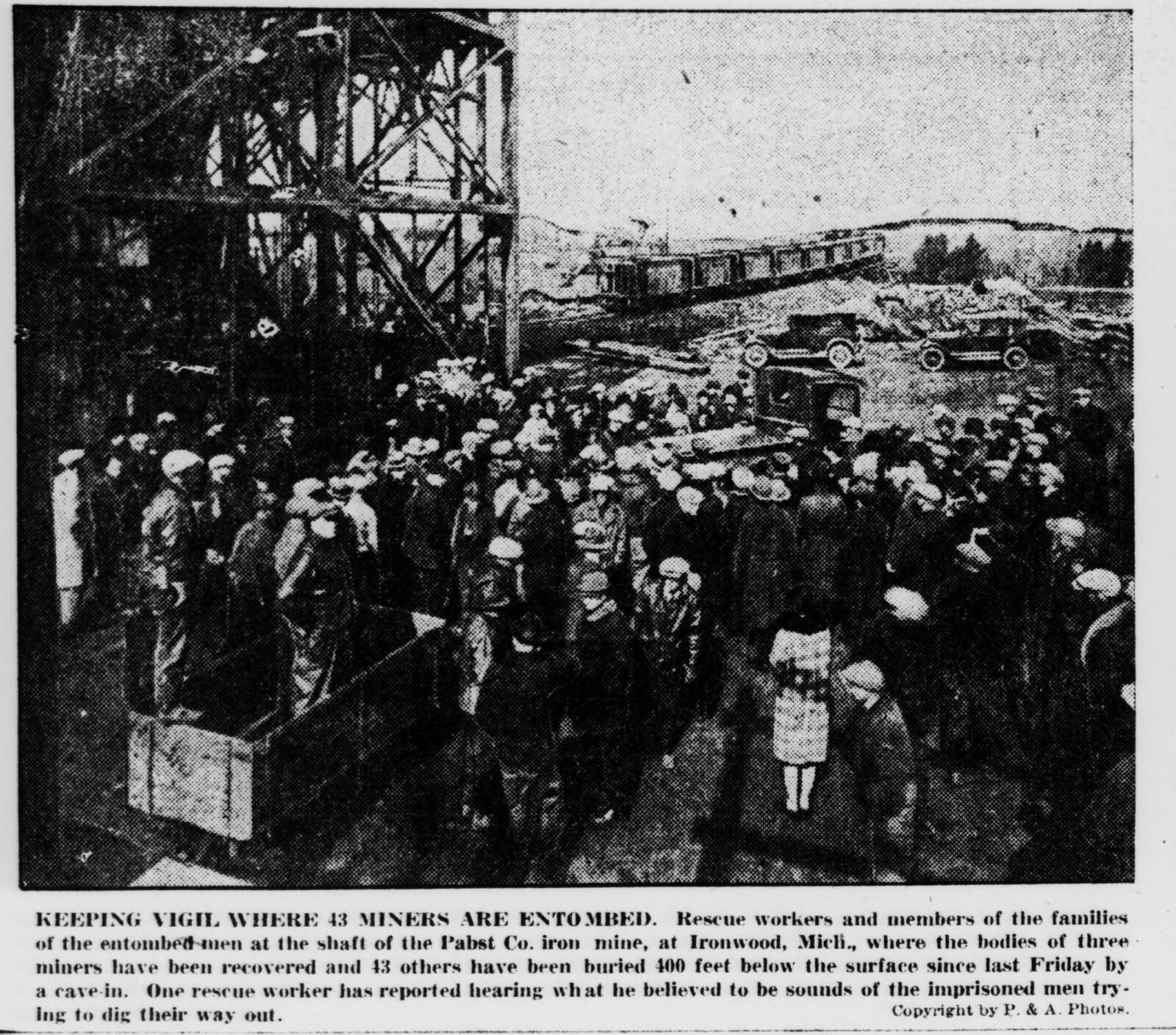 Halftone newspaper photograph: a large crowd in caps and coats stands beneath the steel lattice of a mine headframe; a girl in a pale coat stands at the front, and automobiles and ore cars sit beyond.