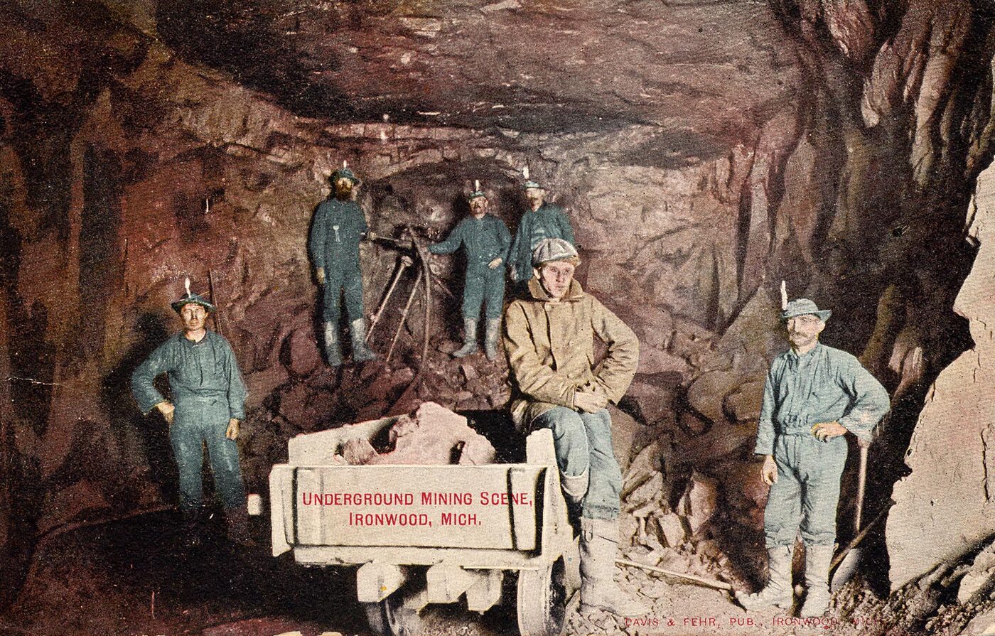 Tinted postcard of an underground chamber in red rock: six miners, several with lamps on their hats, stand around a loaded ore car and a rock drill mounted on a post.