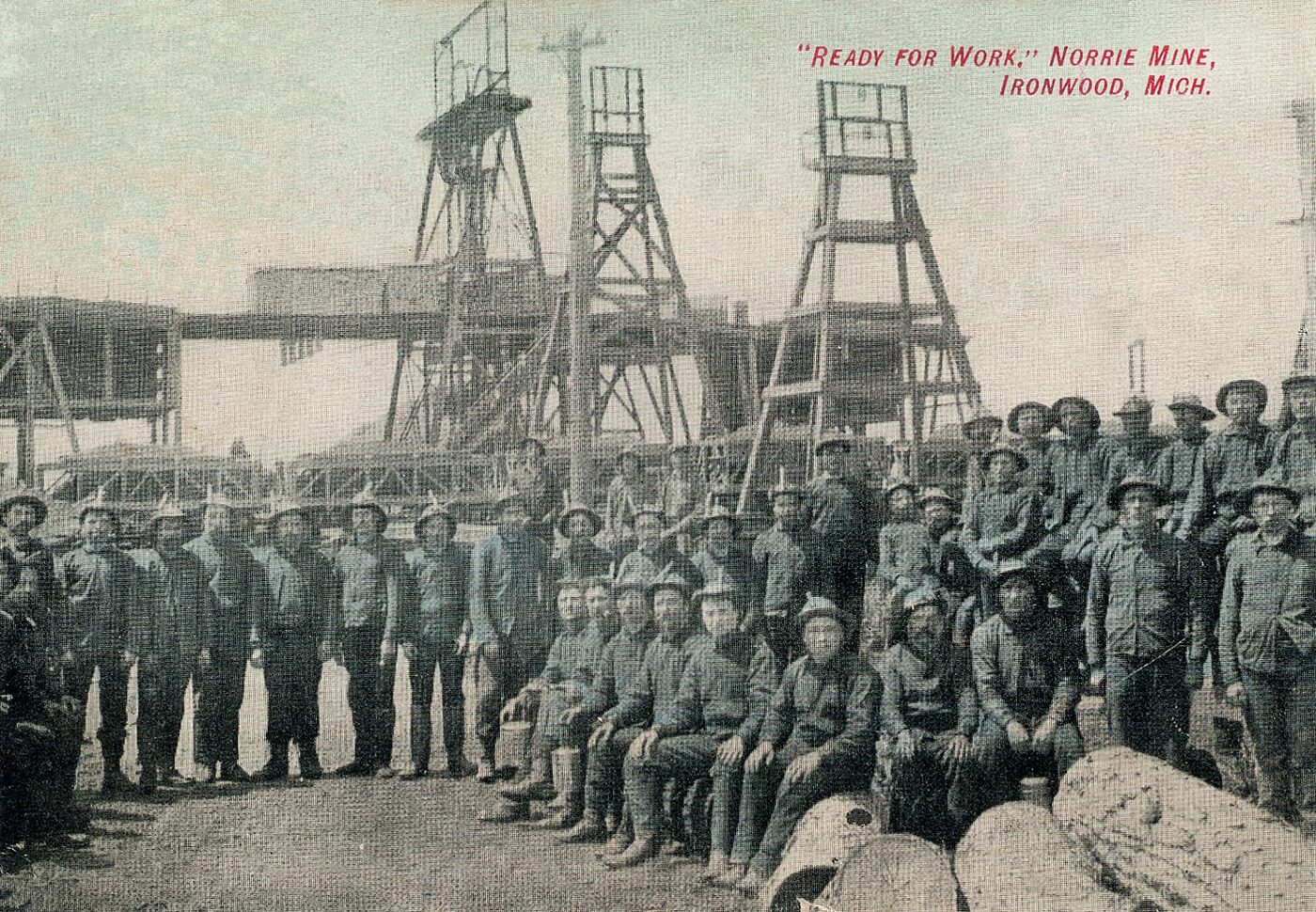 Tinted postcard: several dozen miners in caps and work clothes pose in rows, some seated on logs, before tall steel-lattice headframes at the Norrie Mine, Ironwood.